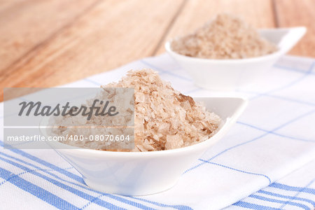 Brown natural salt flakes in white bowl on kitchen cloth on wooden background. Luxurious cooking seasoning.