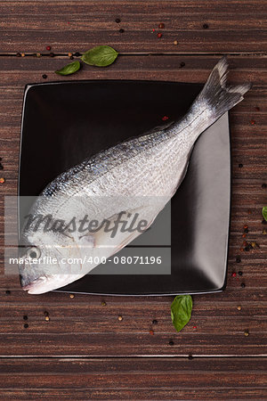 Fresh gilt head bream on black square plate with colorful peppercorn and fresh basil leaves on brown natural background. Culinary seafood concept.