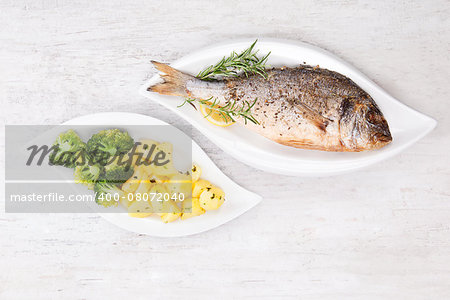 Grilled gilthead bream with fresh rosemary herb, lemon and steamed potatoes and broccoli on white plate on white wooden background. Culinary bright seafood eating.