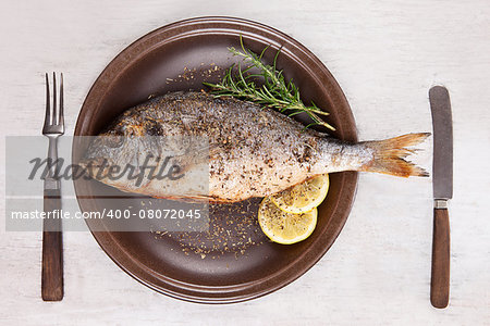 Grilled fish with lemon and rosemary on brown plate with antique cutlery isolated on white wooden background with clipping path, top view. Mediterranean seafood background.