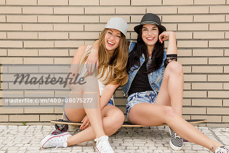 Two beautiful and young girlfriends having fun with a skateboard, in front of a brick wall