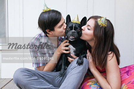 Young couple kissing dog on cheek and wearing party hats