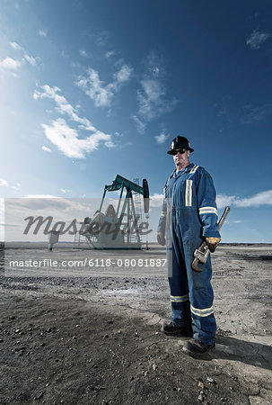 A man in overalls and hard hat at a pump jack in open ground at an oil extraction site.