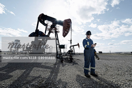 A man in overalls and hard hat at a pump jack in open ground at an oil extraction site.