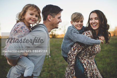Young couple giving son and daughter piggy backs in field