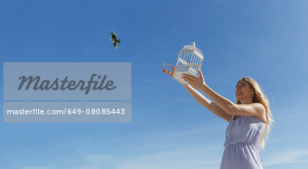 Young woman releasing bird from cage toward blue sky