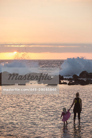 Silhouetted female toddler and mother paddling in sea looking out at waves splashing, Kona, Hawaii, USA