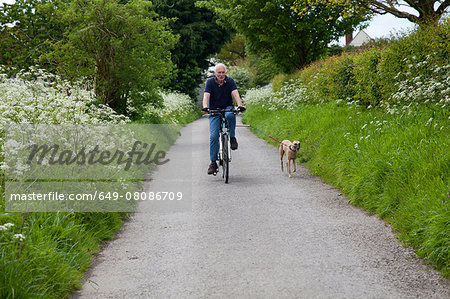 Senior man riding bike on country lane with dog