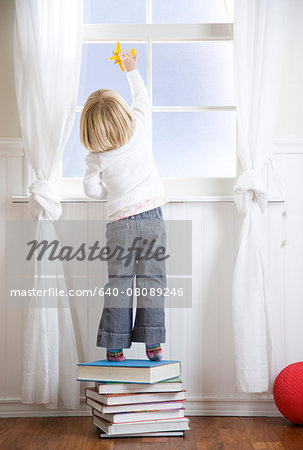 child standing on a stack of books looking out the window