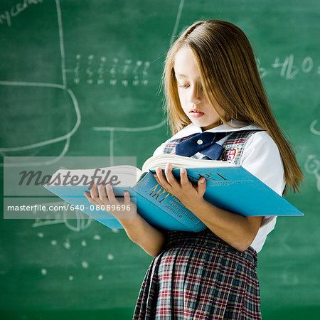 girl in a classroom standing in front of a chalkboard holding a book