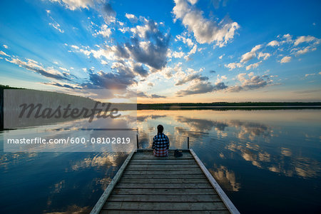 Hiker Sitting on Dock at Calm Lake at Sunset, Saskatchewan, Canada Hiker Sitting on Dock at Calm Lake at Sunset, Saskatchewan, Canada