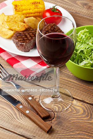 Glass of red wine, steak with grilled potato, corn and salad on wooden table