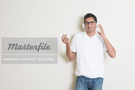 Portrait of handsome Indian guy enjoying music, standing on plain background with shadow, looking at camera.