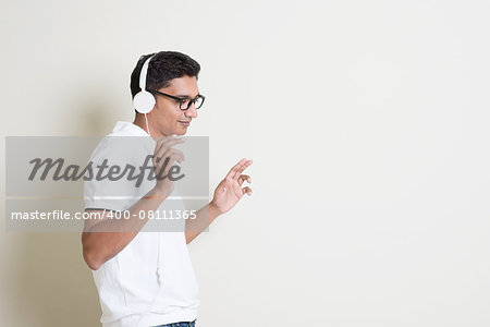 Portrait of handsome Indian guy listening to music with headphones, standing on plain background with shadow, copy space at side.