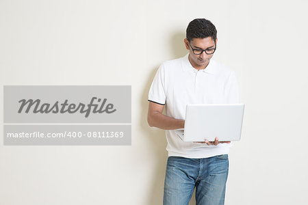 Portrait of handsome Indian guy using notebook computer, standing on plain background with shadow, copy space at side.