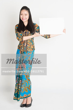 Full body portrait of Southeast Asian girl in batik dress hands holding white blank card, standing on plain background.