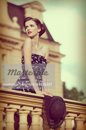 beautiful young brunette girl posing like vintage diva on balcony of ancient palace with elegant hair-style, red lipstick, short dress and hat in the hand .vintage color effect