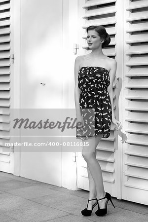 black and white fashion brunette woman posing in summertime in outdoor portrait with sexy short dress, elegant hair-style and black heels.