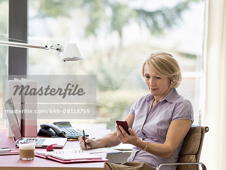 Mature businesswoman reading text message on smartphone at home desk