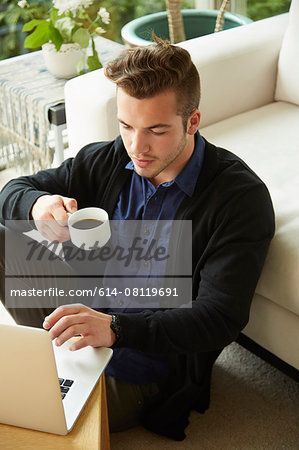 Portrait of man sitting on floor at home looking at laptop and drinking cup of coffee