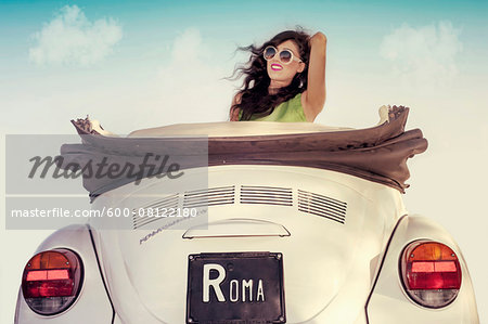 Young woman wearing sunglasses with wind in hair, sitting in a vintage, convertible Volkswagen Beetle, Italy