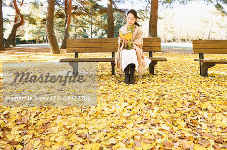 Senior Japanese woman sitting on a bench with a book in a city park in Autumn