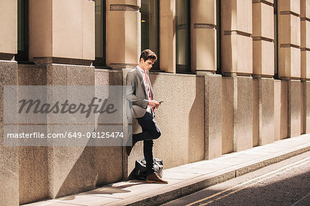 Young city businessman leaning against office building reading smartphone text
