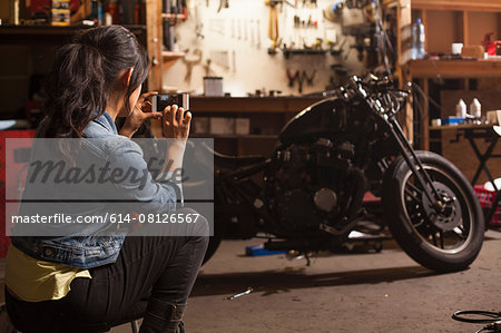 Female mechanic in workshop, taking photograph of motorcycle using smartphone