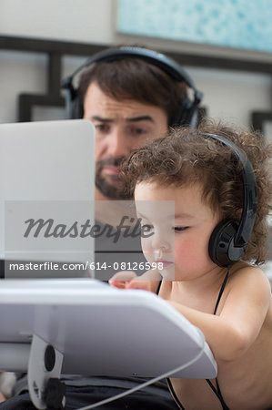 Father watching toddler daughter typing on laptop whilst wearing headphones