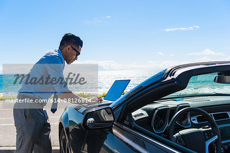 Young businessman at coast parking lot typing on laptop on car hood