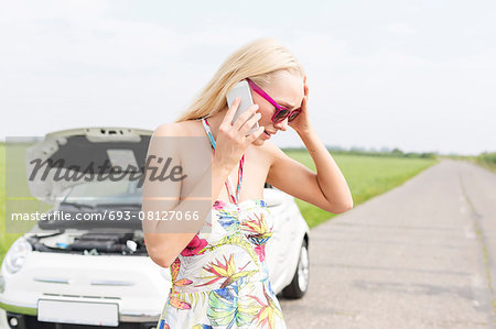 Worried woman using mobile phone on country road with broken down car in background
