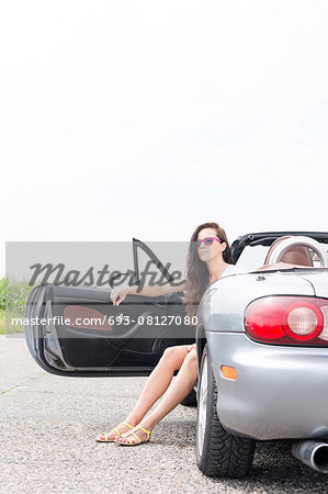 Young woman sitting in convertible on country road against clear sky