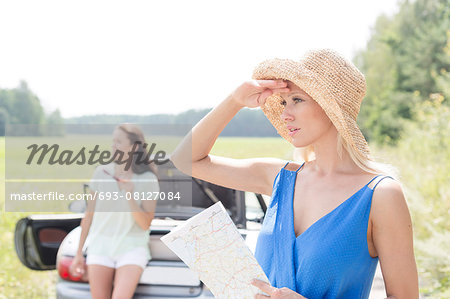 Young woman with map looking away while friend leaning on convertible in background