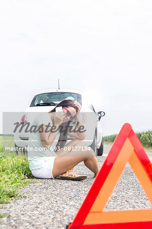 Tensed woman using cell phone while sitting on country road by broken down car