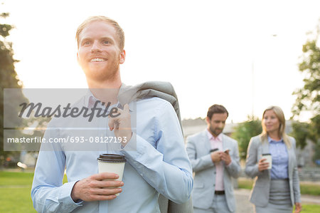 Happy businessman holding disposable cup with colleagues standing in background