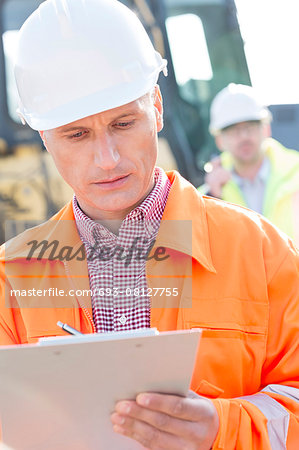 Supervisor writing on clipboard at construction site with colleague in background