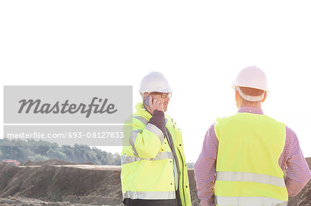 Male engineer using mobile phone while standing with colleague at construction site against clear sky