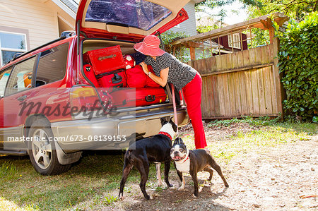 Woman packs luggage into the trunk of a car while her dogs watch