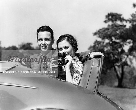 1930s COUPLE SITTING IN JUMP SEAT OF CAR POINTING CAMERA AT VIEWER