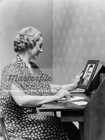 1930s 1940s SENIOR WOMAN SITTING AT DESK WRITING LETTER AS SHE HOLDS PICTURE PORTRAIT OF YOUNG MAN SON