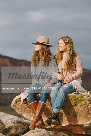 Two women wearing leather boots sitting side by side on a rock in a desert.