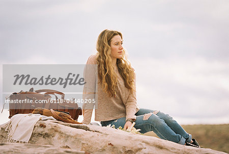 Woman sitting on a rock in a desert, a leather bag standing beside her.