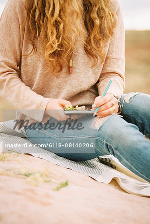 Woman sitting in a desert on a rock, holding a notebook and pencil.