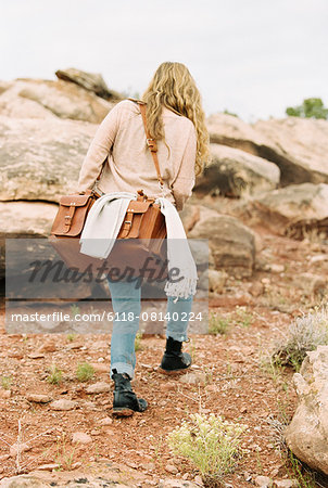 Woman walking past rocks in a desert, carrying a leather bag.