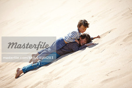 Two young boys lying on sandy hill, playing