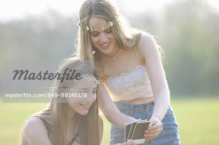 Two teenage girls wearing daisy chain headdresses looking at instant photographs in park