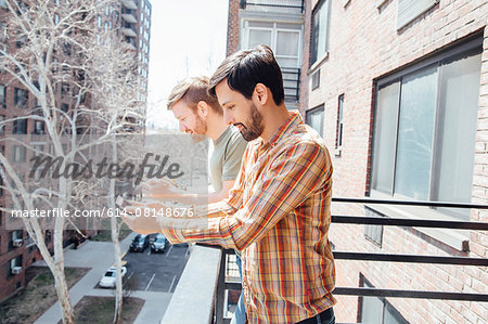 Male couple standing on balcony, looking at smartphones