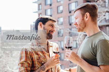 Male couple standing on balcony, holding wine glasses, face to face