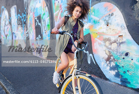 Portrait serious woman with afro on bicycle next to urban graffiti wall Portrait serious woman with afro on bicycle next to urban graffiti wall