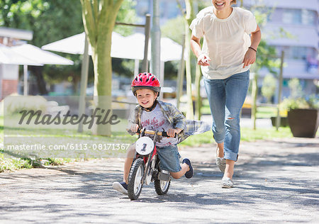 Mother chasing son riding bicycle with helmet in sunny park